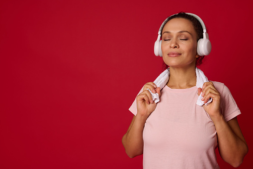 Beautiful middle-aged active Hispanic woman, sportswoman in wireless headphones and pink t-shirt posing with closed eyes on a red background with space for advertising. Sport and fitness concept