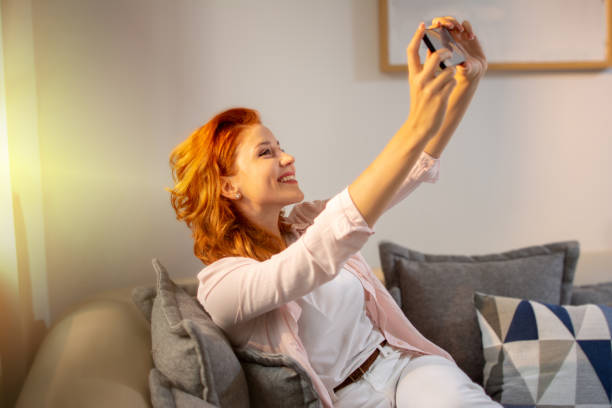 istock Beautiful young red haired woman relaxing on sofa and using smartphone to take a selfie photos for social media at home. 1396524857