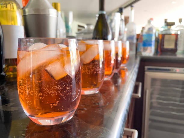 Row of stemless glasses containing orange colored sparkling beverage made of alcohol, liquor, and soda water Close-up of five glasses filled with an orange beverage, ice cubes and bubbles in a row sitting on the edge of a dark colored bar. Glasses are surrounded by bottles of alcohol which are out of focus Soda Water stock pictures, royalty-free photos & images