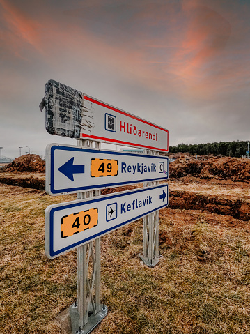 Metal road signs indicating the direction to Hlíðarendi, Reykjavik and Keflavik. Metal signage in forefront with the countryside of Iceland in the background.
