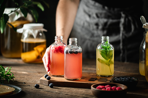 Woman hand serving natural probiotic drink. Female hand placing flavored kombucha tea bottles on wooden table with mint leaves, raspberry and blueberries.