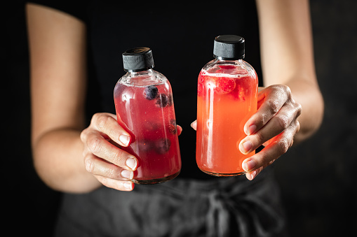 Close-up of woman hands holding two bottles of kombucha tea. Cropped shot of female with raspberry and blueberry fermented tea bottles.