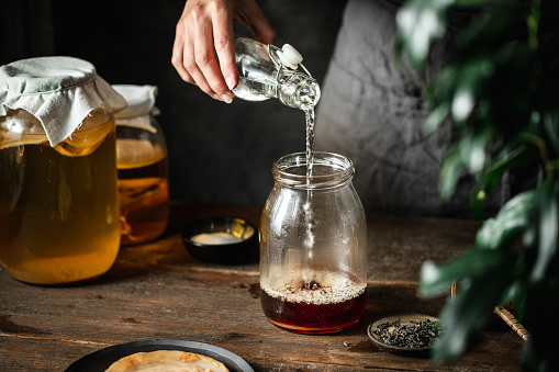 Woman hand pouring water in black tea jar for making kombucha in kitchen. Female making healthy probiotic drink at home.