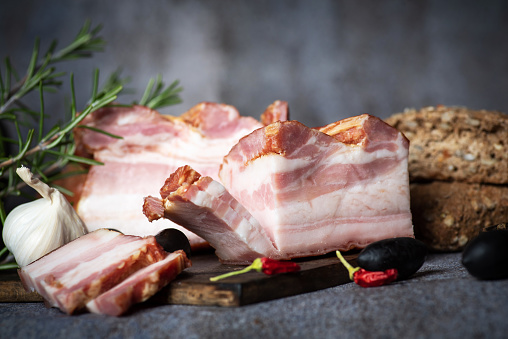 Delicious artisanal whole smoked pork  slab bacon on a cutting block with fresh spices and herbs on dark background