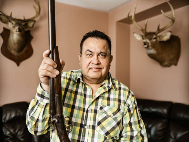 Mature Latin man of Mexican heritage posing with guns and hunting trophies. He is wearing casual clothes with plaid shirt. Interior of private home with taxidermy animal heads on the wall.