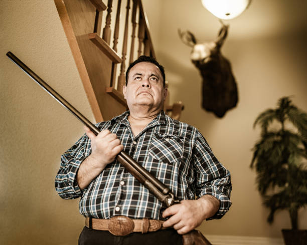 Mature Latin man of Mexican heritage posing with guns and hunting trophies. He is wearing casual clothes with plaid shirt. Interior of private home with taxidermy animal heads on the wall.