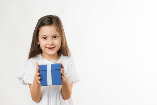 Front view of a cute little girl is holding a blue gift box in her hands with a nice smile in front of pure white background.