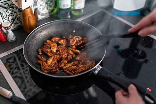 Woman uses a spatula to prepare chicken teriyaki in a pan on the stovetop in the kitchen. Healthy and delicious food at home.