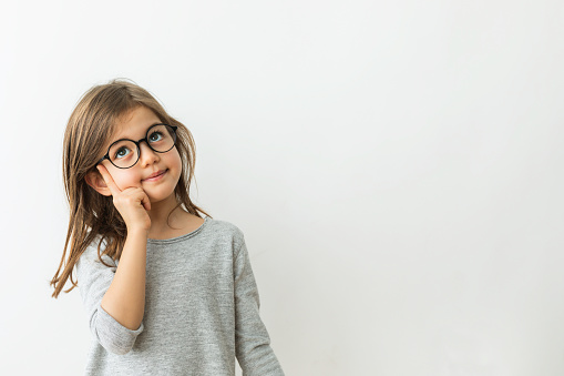 Little girl with eyeglasses is looking up with thinking gesture in front of white background