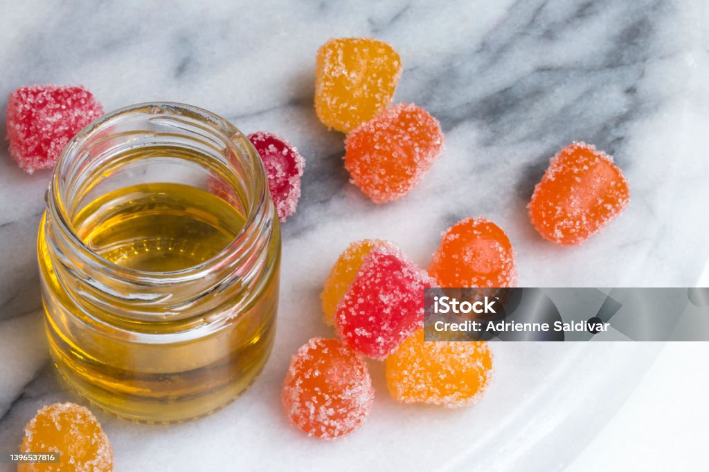 CBD gummy candy gumdrops and oil on a white marble surface CBD gummy candy gumdrops in red, yellow, and orange surrounding a jar of hemp oil on a gray and white marble surface. High angle view. Cannabidiol Stock Photo