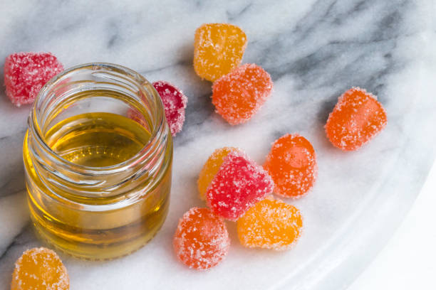 CBD gummy candy gumdrops and oil on a white marble surface CBD gummy candy gumdrops in red, yellow, and orange surrounding a jar of hemp oil on a gray and white marble surface. High angle view. gum drop photos stock pictures, royalty-free photos & images