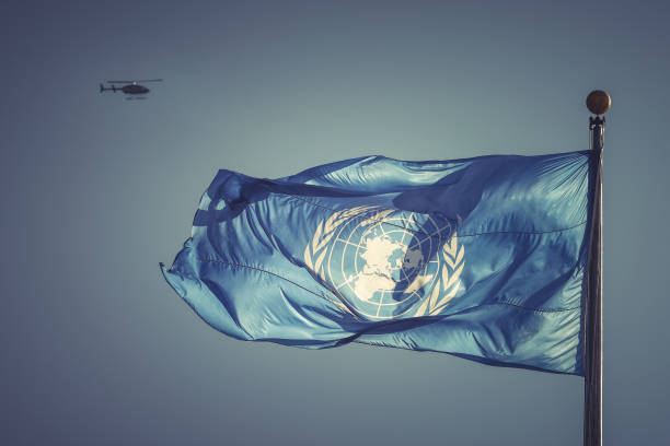 United Nations flag with the UN symbol and a blurred UN helicopter on the background A UN blue flag captured flying in the wind at the United Nations Headquarters with a blurred helicopter on the background symbolizing a dedicated job of UN blue helmets serving in one of UN peacekeeping missions around the globe. Action for peacekeeping concept. united nations stock pictures, royalty-free photos & images