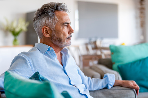 Portrait of a mature man sitting on the sofa at home. He is serious looking away from the camera.