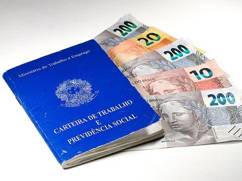Translation: 'Federative Republic of Brazil, Ministry of Labor'. Brazilian Work Card (Carteira de Trabalho) with Brazilian money on white background. Brazilian economy concept.