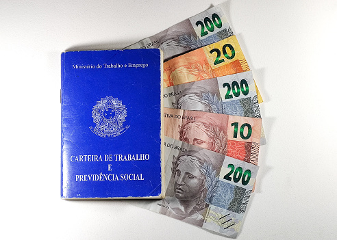 Translation: 'Federative Republic of Brazil, Ministry of Labor'. Brazilian Work Card (Carteira de Trabalho) with Brazilian money on white background. Brazilian economy concept