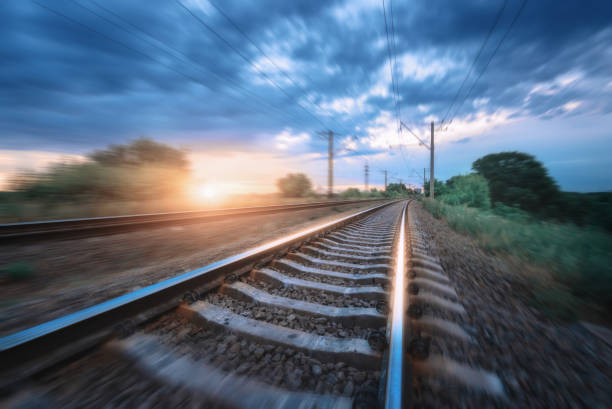 istock Railroad and dramatic cloudy blue sky at sunset with motion blur effect in summer. Railway station in motion. Industrial landscape with blurred railway platform, green trees and grass in the evening 1396560324