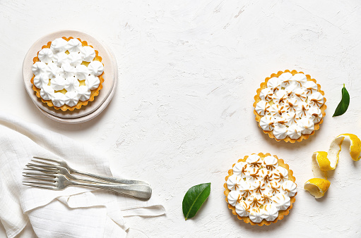 Mini lemon pies with meringue, lemon peel, lemon leaves, forks, and a white napkin, on a white background with copy space.