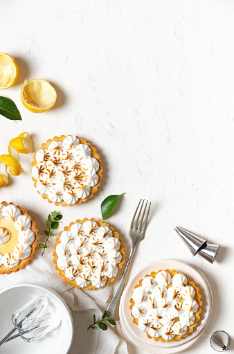 Mini lemon pies with meringue, lemon peel, lemon leaves, whisks with meringue, a fork, and some piping nozzles, on a white background with copy space.