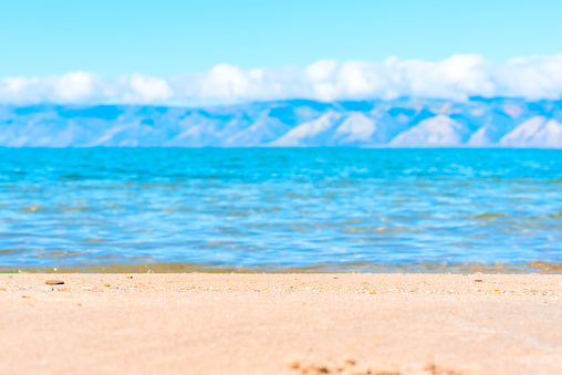 Narrow strip of focused sand on the beach with defocused water of Lake Baikal and mountains at sunny summer day. Beauty of nature concept, travel background.