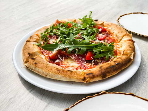 Side view of pizza with prosciutto, arugula, tomatoes, and parmesan on a white plate on the table. Italian cuisine. Stand. Dough. Nutrition. Menu. Circle. Small. Slice. Prosciutto. Sauce. Dish
