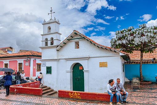 Bogota, Colombia - July 20, 2016: The Ermita or Hermitage of San Miguel del Principe on the Chorro de Quevedo in the South American, Andean capital city of Bogota, in Colombia. The chapel was built in 1969 based on images of the Capilla del Humilladero which is said to have been the first chapel in the New Kingdom of Granada in the early 16th Century. Dark clouds are setting in from one side: it will rain shortly. Some local Colombian people can be seen in the image. Photo shot on a partly cloudy morning; horizontal format. Copy space.