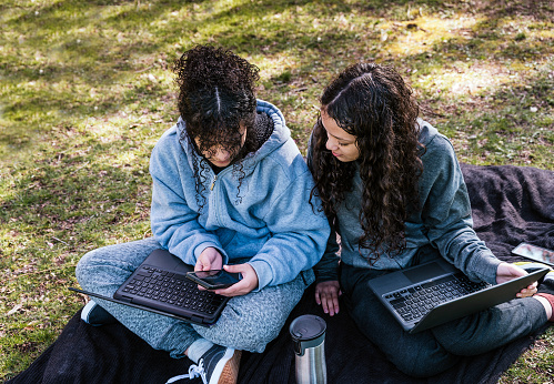 Portrait of cheerful teen sisters, girls looking at mobile phone and computer while sitting together at the park.