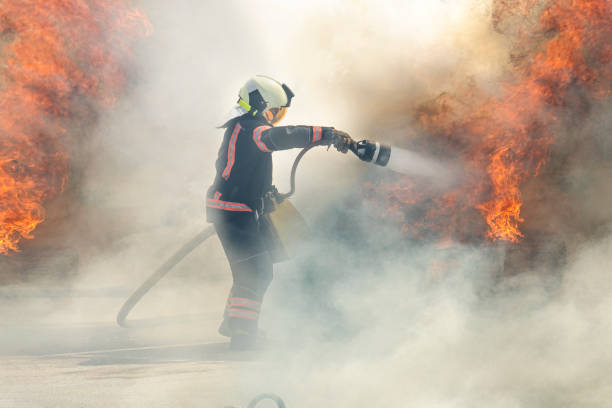 der heldenhafte feuerwehrmann steht zwischen rauch und feuer und löscht das feuer mit einem wasserstrahl. feuerwehrmann löscht ein feuer - löschpapier stock-fotos und bilder