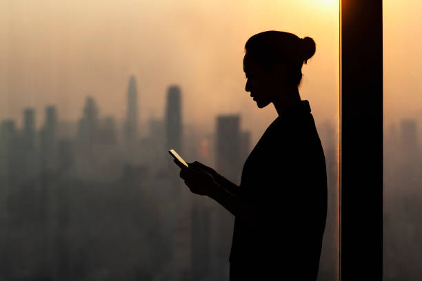 silueta de mujer joven usando smartphone junto a ventana con paisaje urbano - encryption fotografías e imágenes de stock