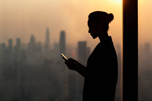 Silhouette of young woman using smartphone next to window with cityscape Silhouette of young woman using smartphone next to window with cityscape, Shenzhen, China anonymous phone stock pictures, royalty-free photos & images