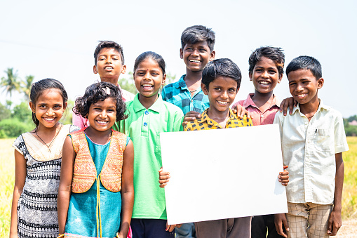 Group of happy smiling Indian village children with one kid holding empty sign board or placard by looking camera - concept of happiness, advertisement and promotion