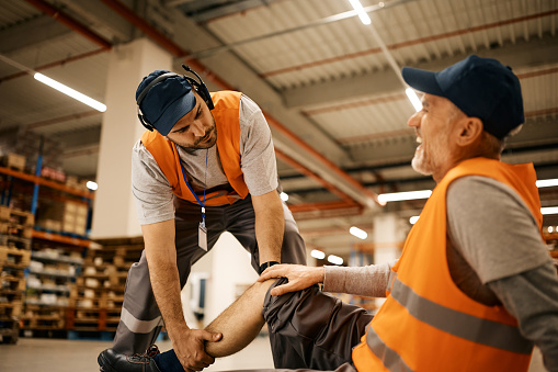Young worker assisting his colleague with leg injury while working at distribution warehouse.