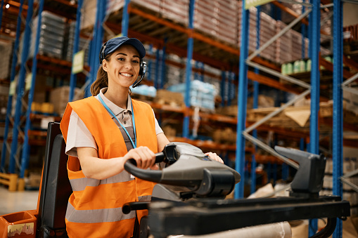 Happy woman working as pallet jack driver at distribution warehouse and looking at camera.