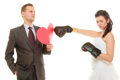 Conflict in relationship concept. Married couple fighting with each other. Woman wearing wedding dress and boxing gloves punching her husband in suit holding heart.