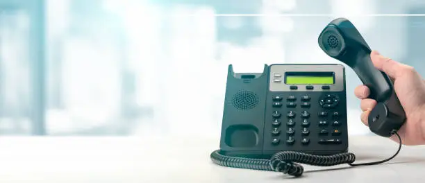 Photo of Dialing telephone keypad concept for communication, contact us and customer service support. black office voip phone with handset up on table on blurred office background. banner