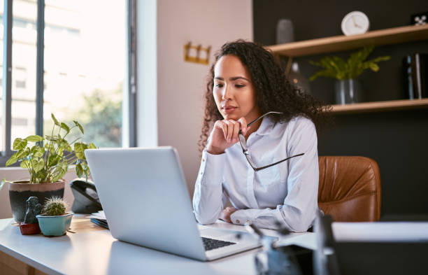 Young and ambitious mixed race entrepreneur sitting alone and thinking while browsing the internet on a laptop in an office. Hispanic business woman using a computer to plan her freelancing startup