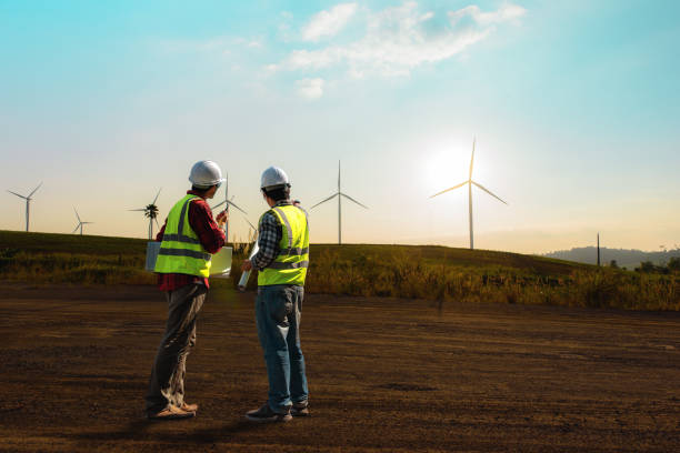 istock Two maintenance engineer working on wind turbine farm in sunset, discussed plan for the maintenance of wind turbines while holding blueprint of layout plan. 1396617344