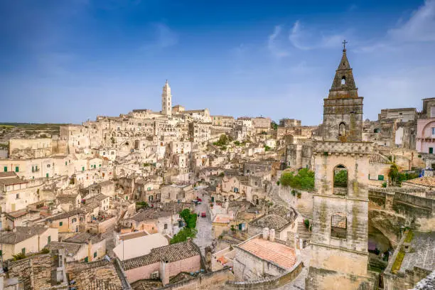 An idyllic cityscape of the old town of Matera, in southern Italy, known worldwide as 'Sassi di Matera' (Matera's Stones). On the horizon stands the majestic facade and the bell tower of the Duomo, or Cathedral of the Madonna della Bruna and of Sant'Eustachio, built in the Apulian Romanesque style in the 13th century. The ancient city of Matera, in the region of Basilicata, is one of the oldest urban settlements in the world, with a human presence that dates back to more than 9,000 years ago, in the Paleolithic period. The Matera settlement stands on two rocky limestone hills called 'Sassi' (Sasso Caveoso and Sasso Barisano), where the first human communities lived in the caves of the area. The rock cavities have served over the centuries as a primitive dwelling, foundations and material for the construction of houses, roads and beautiful churches, making Matera a unique city in the world. In 1993 the Sassi of Matera were declared a World Heritage Site by Unesco. Wide angle image in High Definition format.