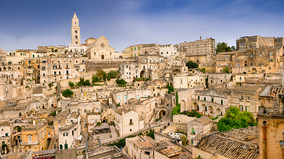 An idyllic cityscape of the old town of Matera, in southern Italy, known worldwide as 'Sassi di Matera' (Matera's Stones). At the center stands the majestic facade and the bell tower of the Duomo, or Cathedral of the Madonna della Bruna and of Sant'Eustachio, built in the Apulian Romanesque style in the 13th century. The ancient city of Matera, in the region of Basilicata, is one of the oldest urban settlements in the world, with a human presence that dates back to more than 9,000 years ago, in the Paleolithic period. The Matera settlement stands on two rocky limestone hills called 'Sassi' (Sasso Caveoso and Sasso Barisano), where the first human communities lived in the caves of the area. The rock cavities have served over the centuries as a primitive dwelling, foundations and material for the construction of houses, roads and beautiful churches, making Matera a unique city in the world. In 1993 the Sassi of Matera were declared a World Heritage Site by Unesco. Super wide angle image in 16:9 and High Definition format.