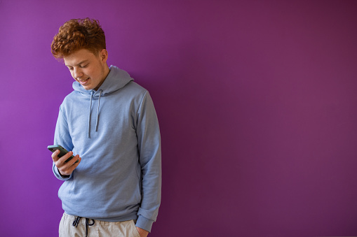 Portrait of Caucasian redhead teenage boy using mobile phone. Purple background