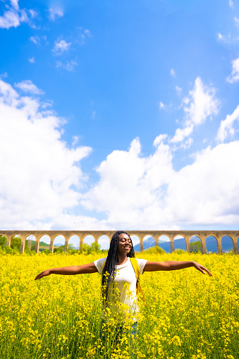 Enjoying spring on vacation, black ethnic girl with braids, traveler, in a field of yellow flowers, vertical photo