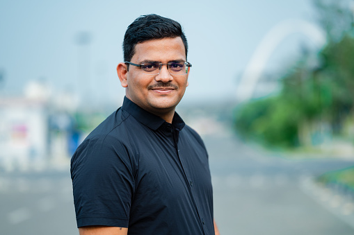 Outdoor portrait image of a happy young man of Indian origin wearing black shirt and looking at camera.