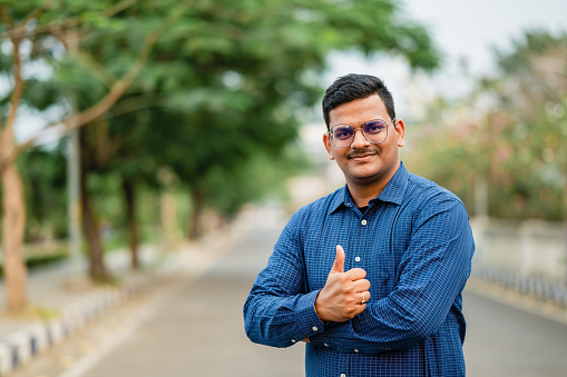 Outdoor image of young Indian man doing positive gesture with hand, thumbs up smiling and happy for success.