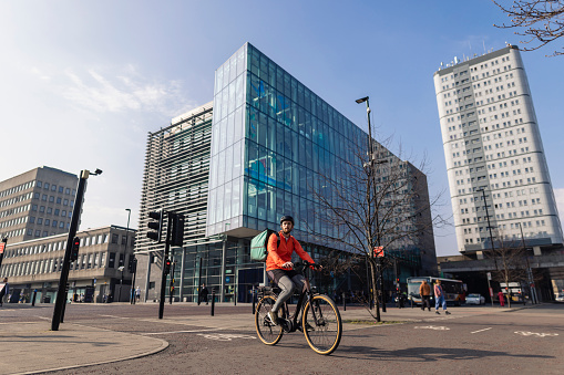 A delivery man wearing a bicycle helmet and food delivery bag, riding his bike on a bike path in Newcastle upon Tyne, England with modern buildings behind him. He is delivering food to a customer.