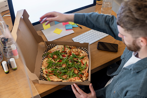 A young man sitting at his desk in his office, he has just opened a cardboard pizza box with a vegan pizza in it after it has been delivered  to him at work.