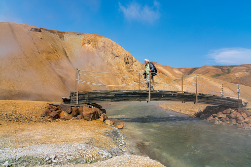 Senior man crossing the bridge over hot spring water at Hveradalir geothermal area on Kerlingarfjoll mountain range, against clear blue sky, Iceland