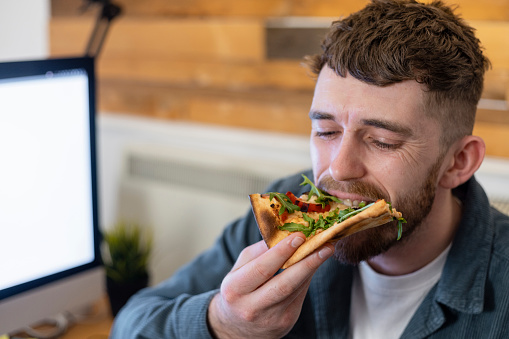 A young man sitting at his desk in an office, eating a slice of vegan pizza that has just been delivered to him at work by a food delivery company.