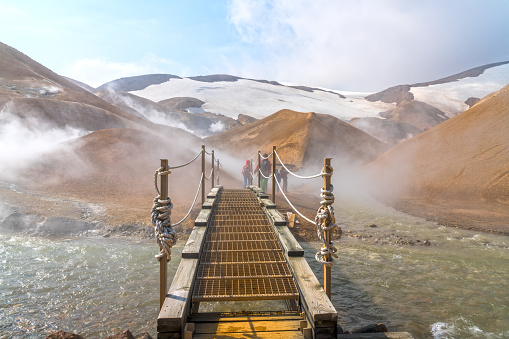 Two tourists hidden in steam crossing the bridge over hot spring water at Hveradalir geothermal area on Kerlingarfjoll mountain range, low angle view, Iceland