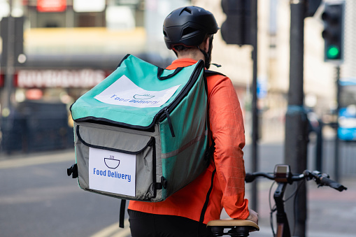 A delivery man wearing a bicycle helmet, walking along a city street while pushing his electric bike along the pavement in Newcastle upon Tyne, England. He has a food delivery backpack on and is off to deliver food to a customer.