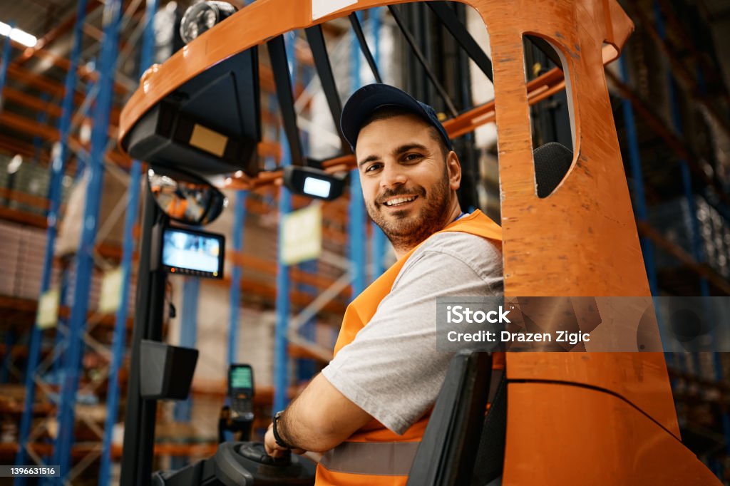 Happy male worker driving forklift at distribution warehouse and looking at camera. Happy forklift operator working at storage compartment and looking at camera. Forklift Stock Photo