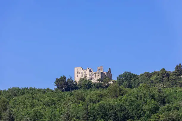 Photo of Intimidating ruins of Lacoste castle, also known as castle of Marquis de Sade, on hill among dense foliage of surrounding forest against clear blue sky. Vaucluse, Provence, France